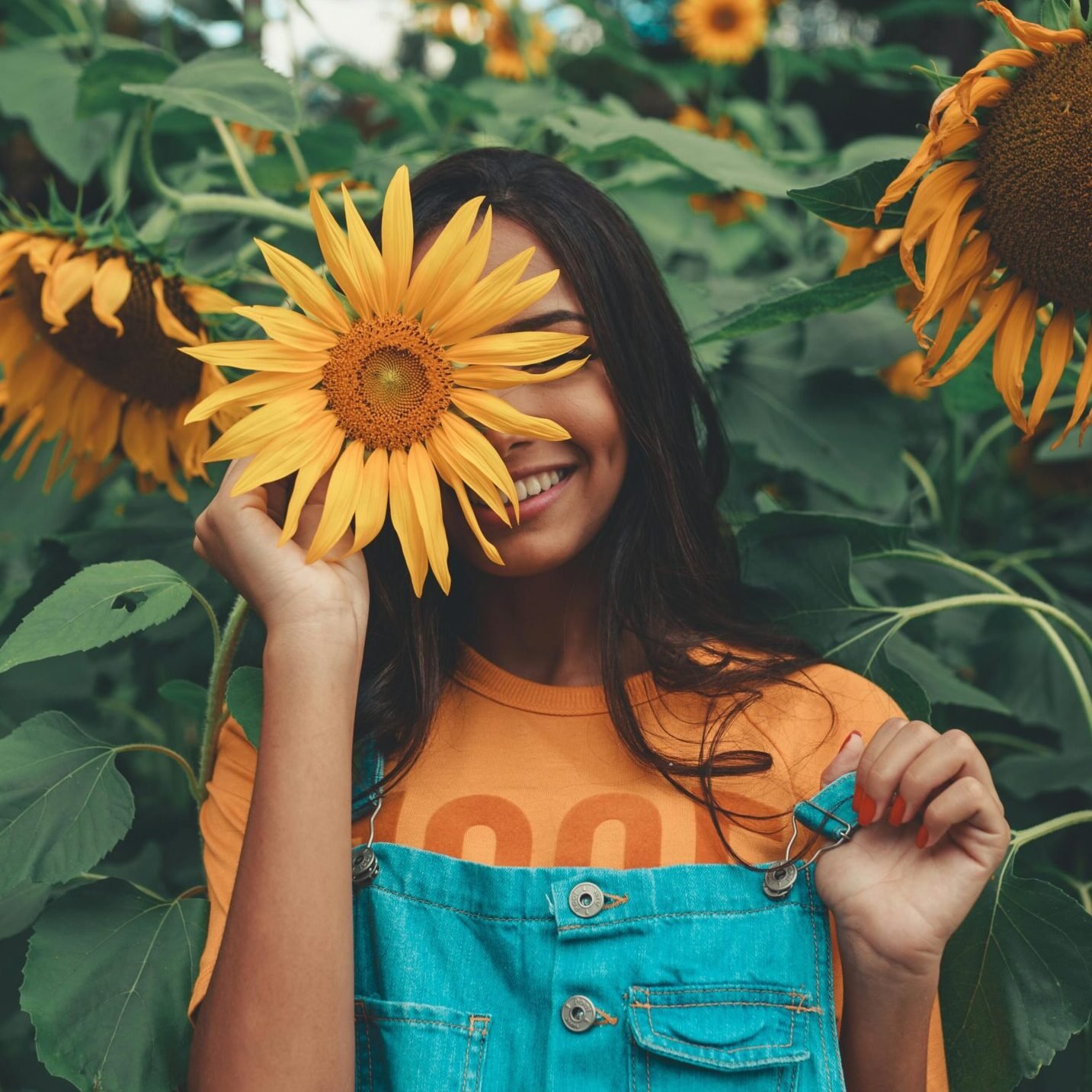 Joyful woman in denim holding sunflower among vibrant blooms in summer garden.